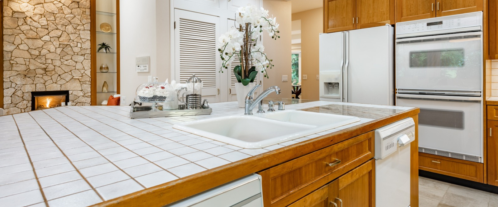 Modern kitchen with white tile countertops, built-in sink, white appliances, wooden cabinets, and a decorative stone fireplace in the background.