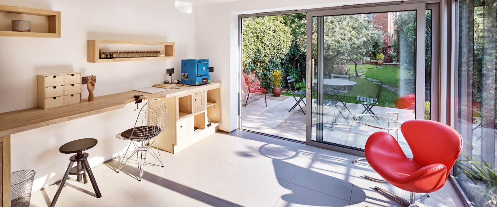 Modern home office with wooden desk, shelves, and a red chair, next to glass sliding doors opening to a garden patio with outdoor seating.