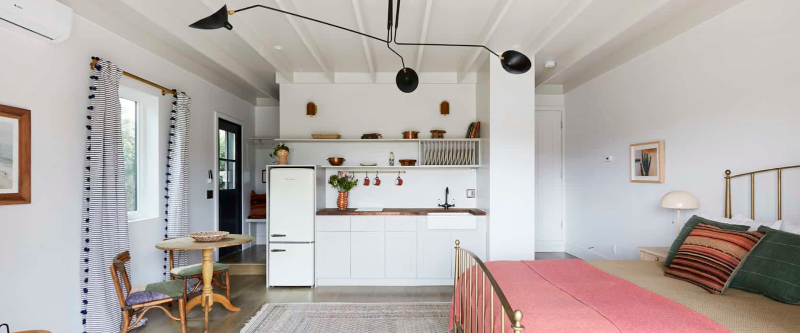 Bright, modern studio with a small kitchen, round dining table, and bed; minimalist decor with white walls, wood accents, and black light fixture.