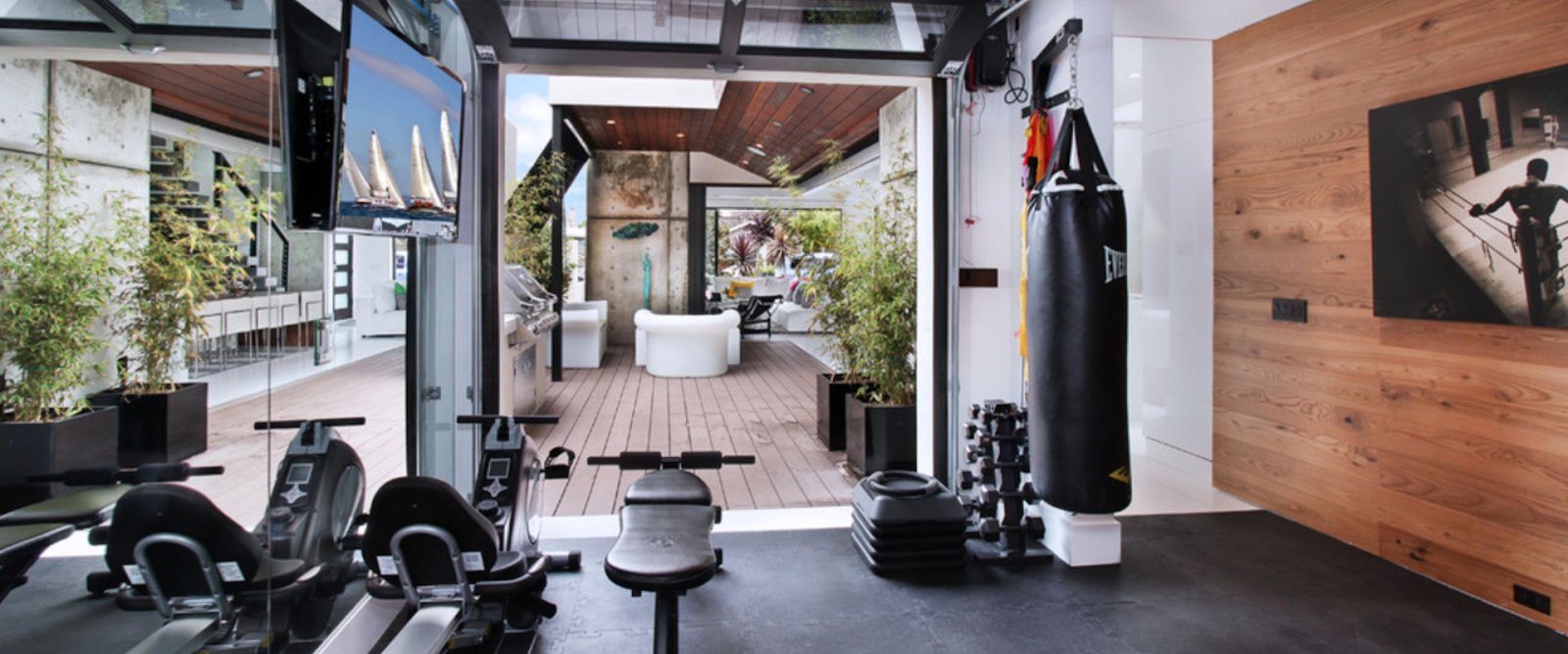 Modern home gym with exercise equipment, punching bag, weights, and a wall-mounted TV; glass doors open to a patio with outdoor seating and greenery.
