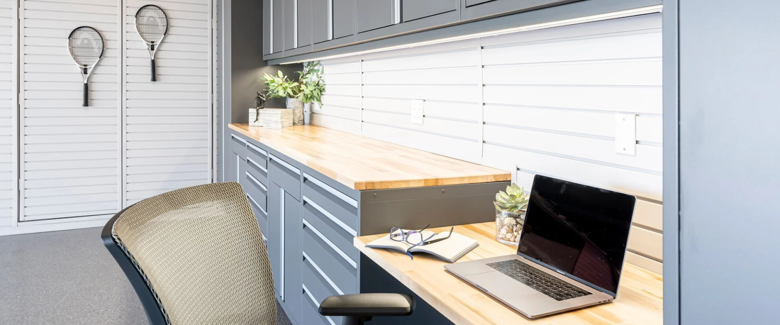 A modern workspace with a laptop, notebook, glasses, and small plants on a wooden countertop, gray cabinets, and two tennis rackets hanging on the wall in the background.