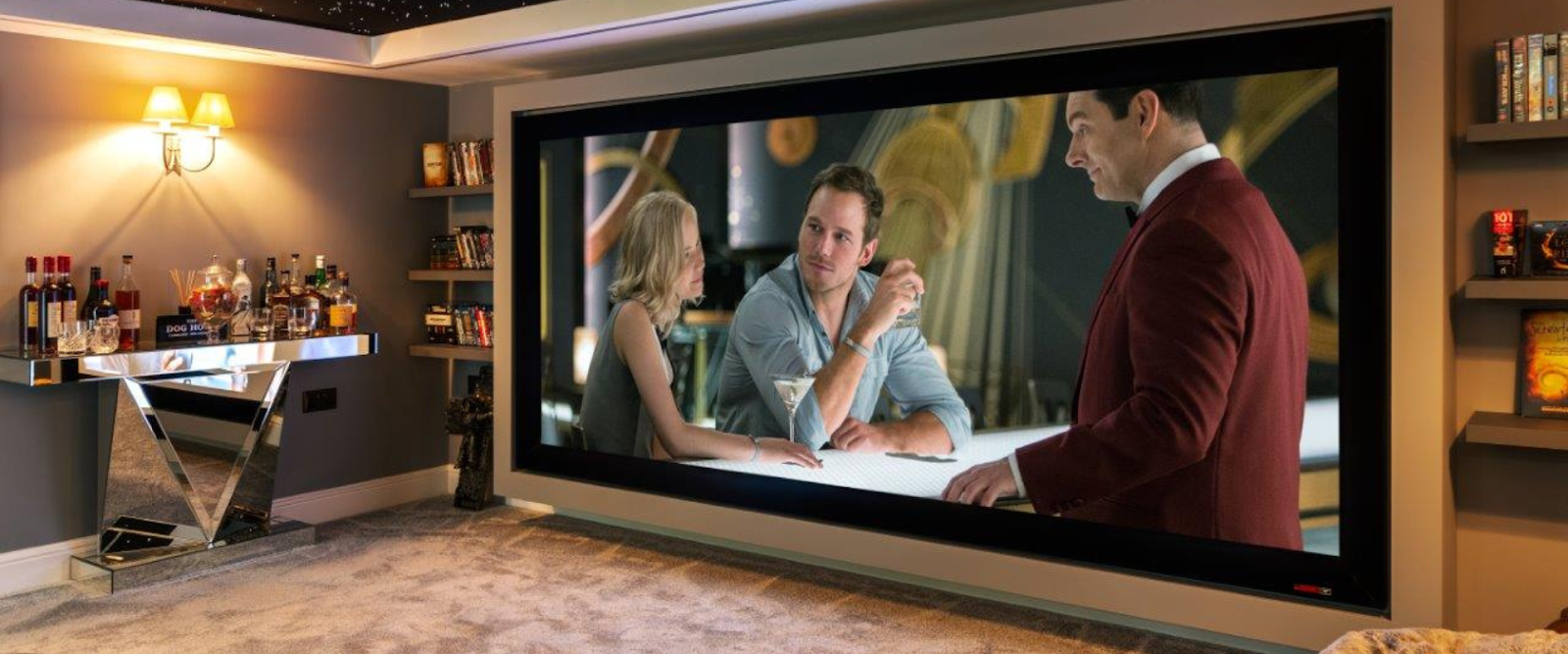 A home theater room with a large screen displaying a movie scene; a bar with various bottles is visible on the left side of the room.