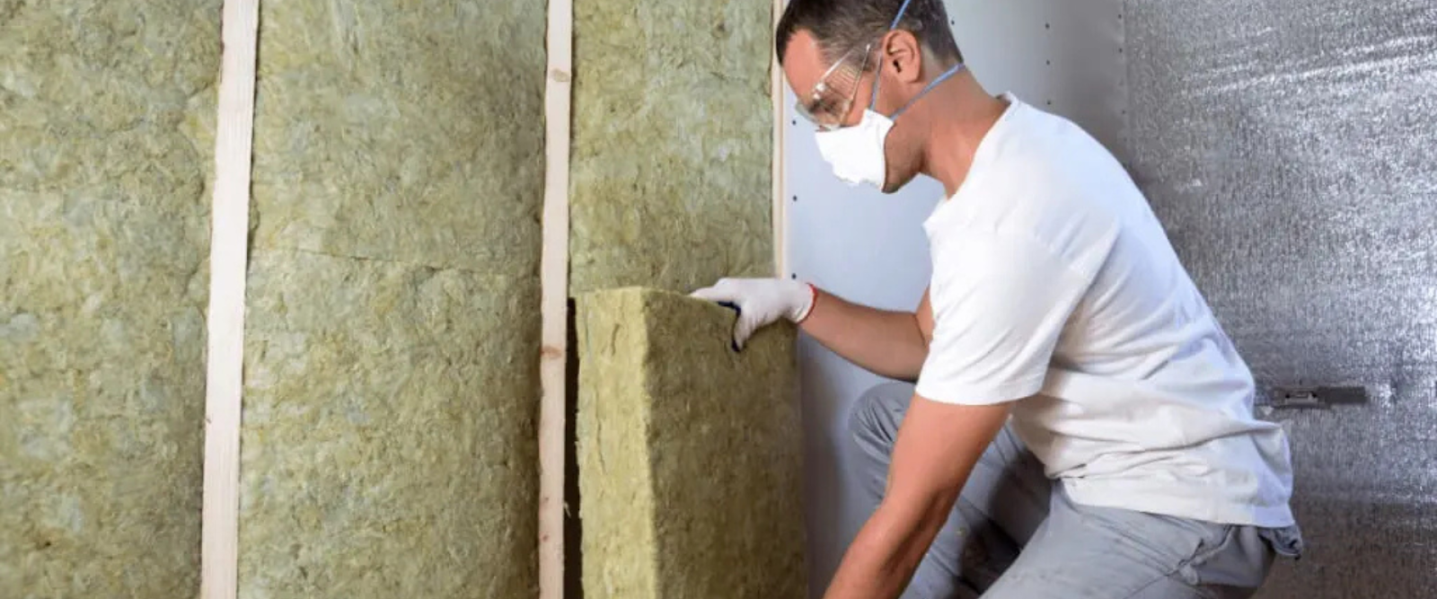 A person wearing protective gear installs fiberglass insulation panels between wall studs in a building under construction.