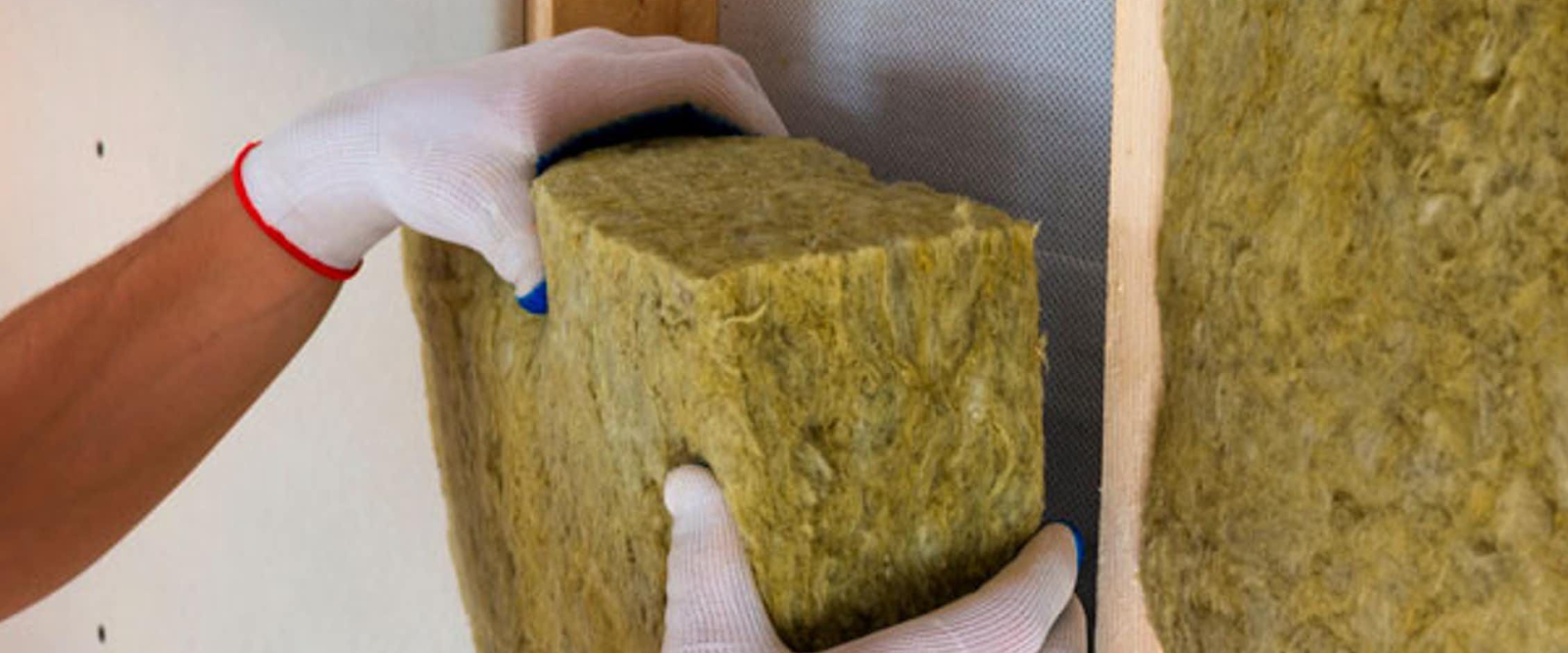 A person wearing white gloves installs a piece of yellow-green insulation material between wooden wall studs.