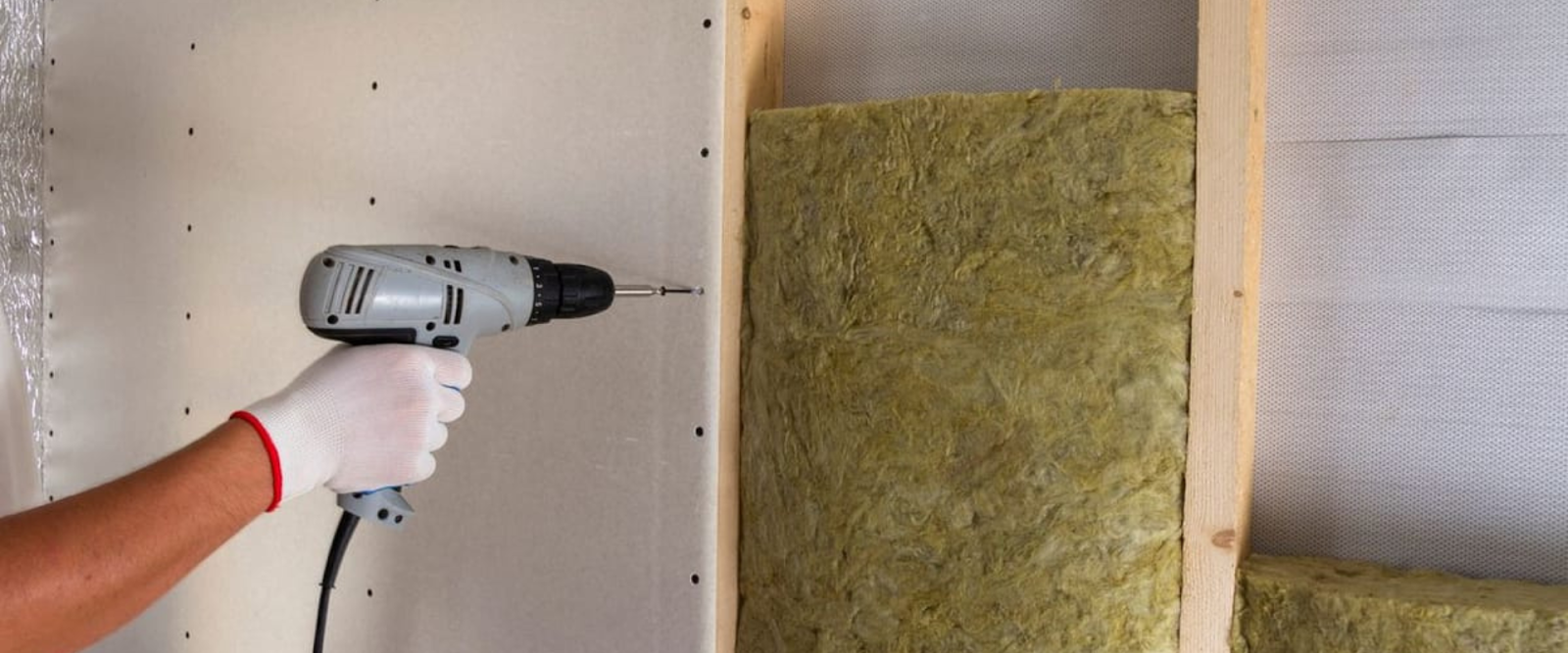 A hand wearing a white glove uses a power drill to install drywall over wall insulation and wooden studs.