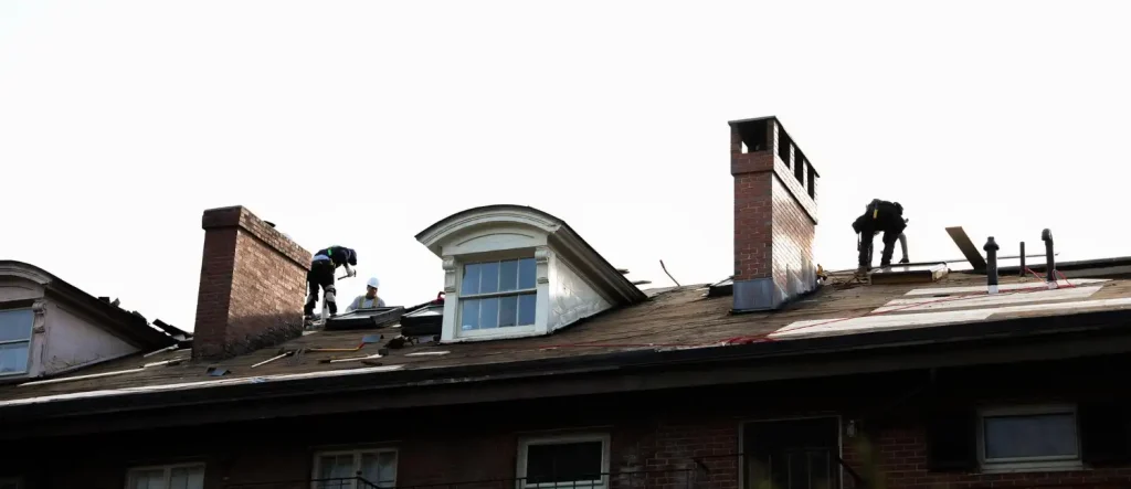Workers repair the roof of a building, with visible tools and materials, near chimneys and a dormer window under a bright sky.