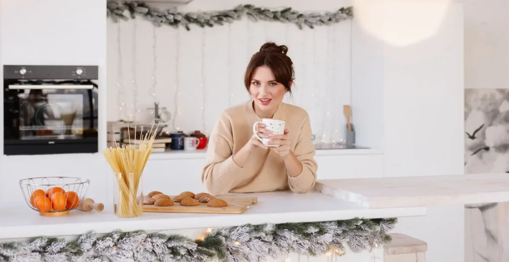 A woman in a beige sweater holds a mug and sits at a kitchen counter with cookies, uncooked spaghetti, and oranges. The kitchen is decorated with holiday garland and lights.