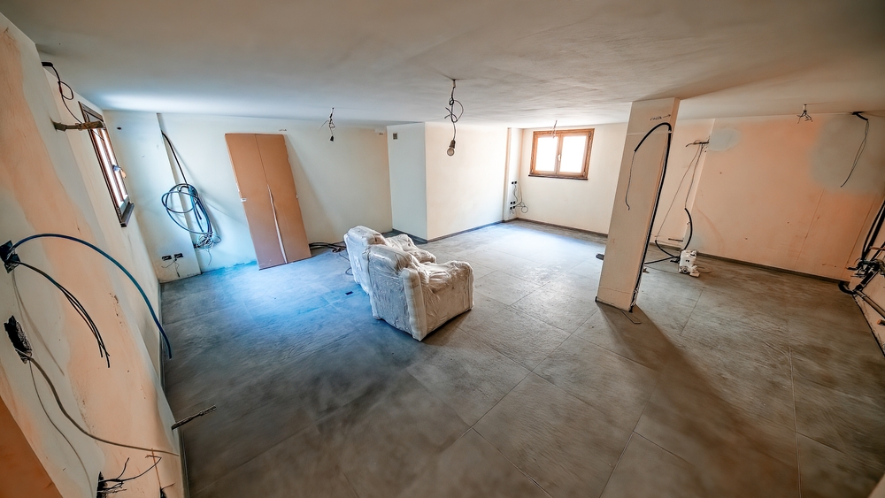Large unfinished room with bare walls, exposed wires, concrete floor, a window, and two wrapped armchairs in the center—perfect for exploring basement finishing ideas to transform this space.