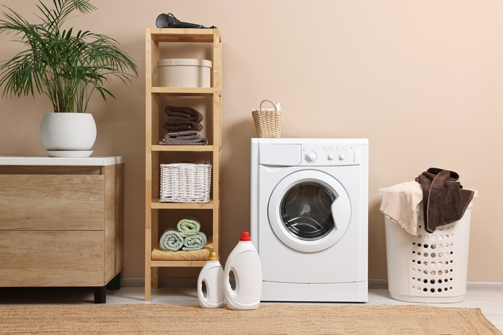 A laundry room makeover idea featuring a washing machine, detergent bottles, a wooden shelf with towels, baskets, a laundry hamper filled with clothes, a plant, and a stylish wooden cabinet.