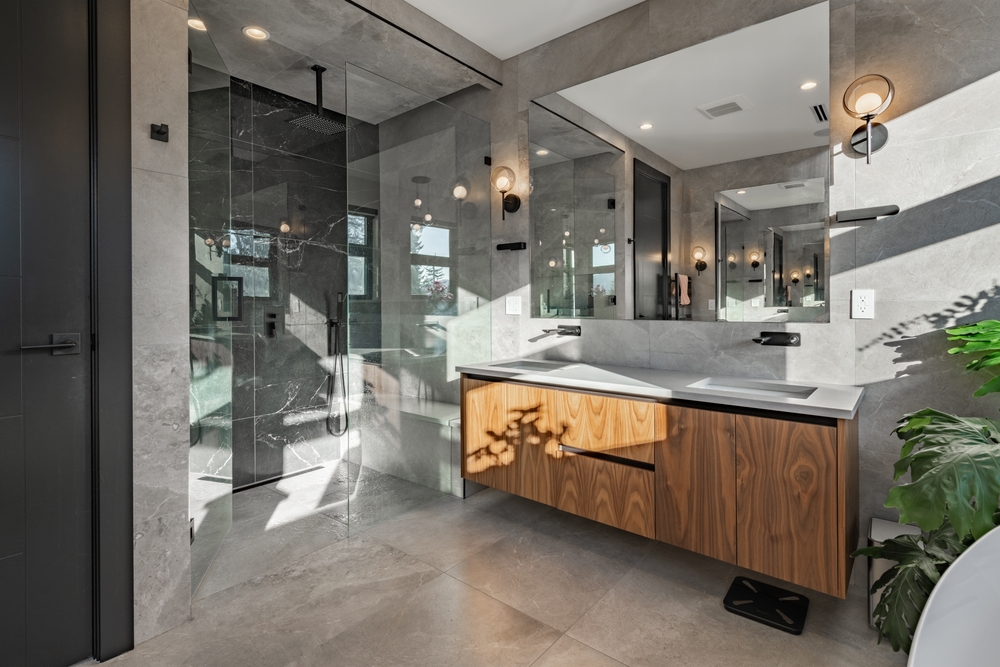 Modern bathroom with gray tile walls and floors, a glass-enclosed shower, wood vanity with a white countertop, large mirrors, wall lights, and a potted plant.